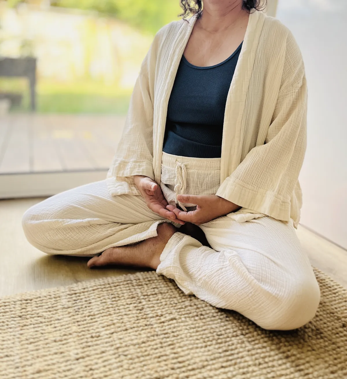 Une femme assise en tailleur sur un tapis de jute, les mains ouvertes sur les genoux, dans la lumière du jour.